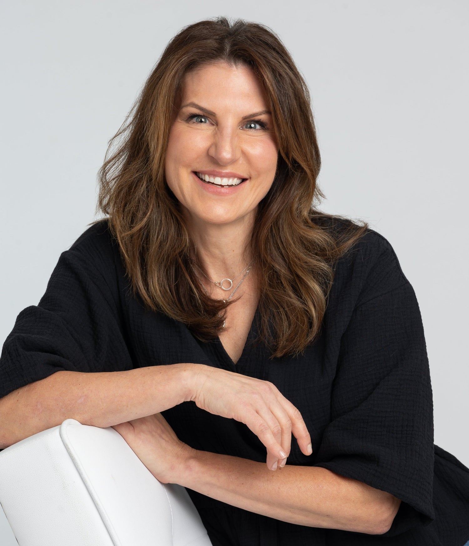 A portrait of Liz Martin, the founder of MYNE Beauty, against a white backdrop wearing a black shirt and sitting on a white chair.