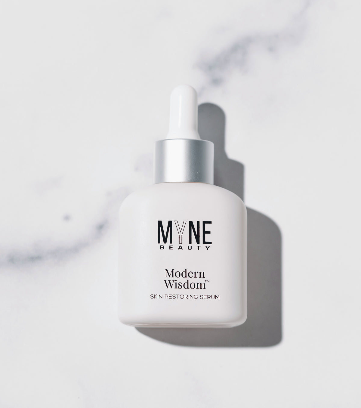 A small, white bottle of Modern Wisdom skin restoring serum from Myne Beauty laid down on a white marble countertop., t4