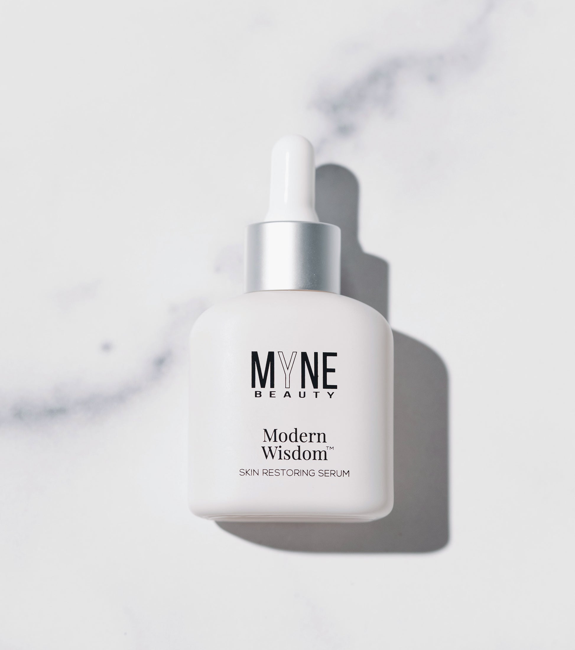 A small, white bottle of Modern Wisdom skin restoring serum from Myne Beauty laid down on a white marble countertop.