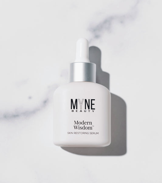 A small, white bottle of Modern Wisdom skin restoring serum from Myne Beauty laid down on a white marble countertop.