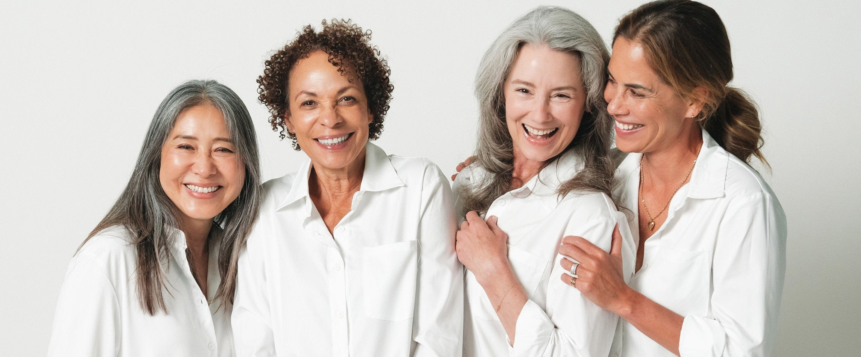 Four women wearing white long sleeve shirts posing for a group photo in front of a white wall.