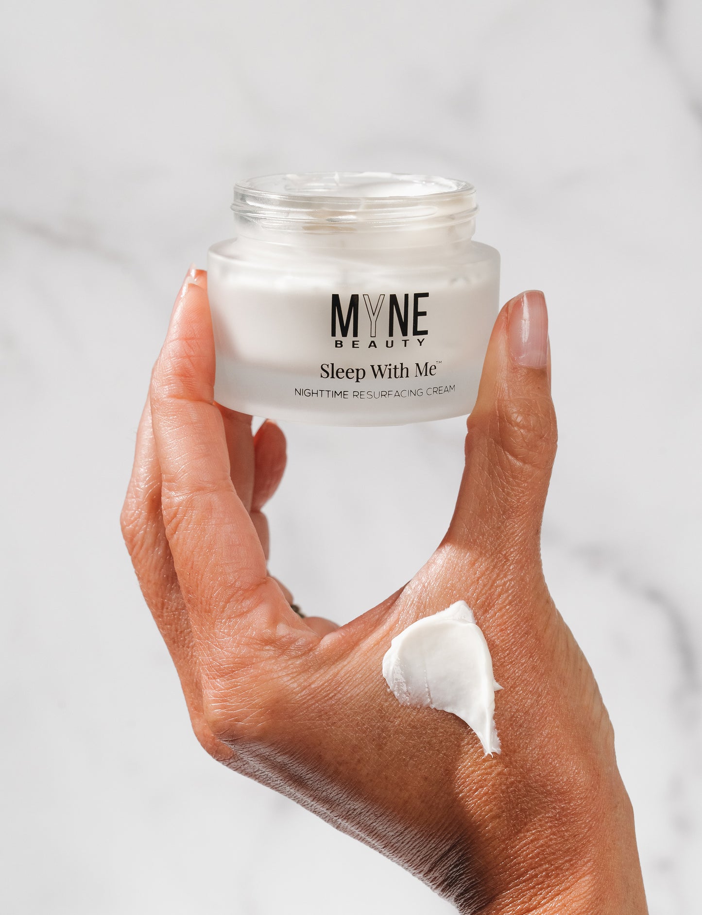 A person holding a jar of Sleep With Me Nighttime Resurfacing Cream between their fingers, with a small dab smeared on their wrist.